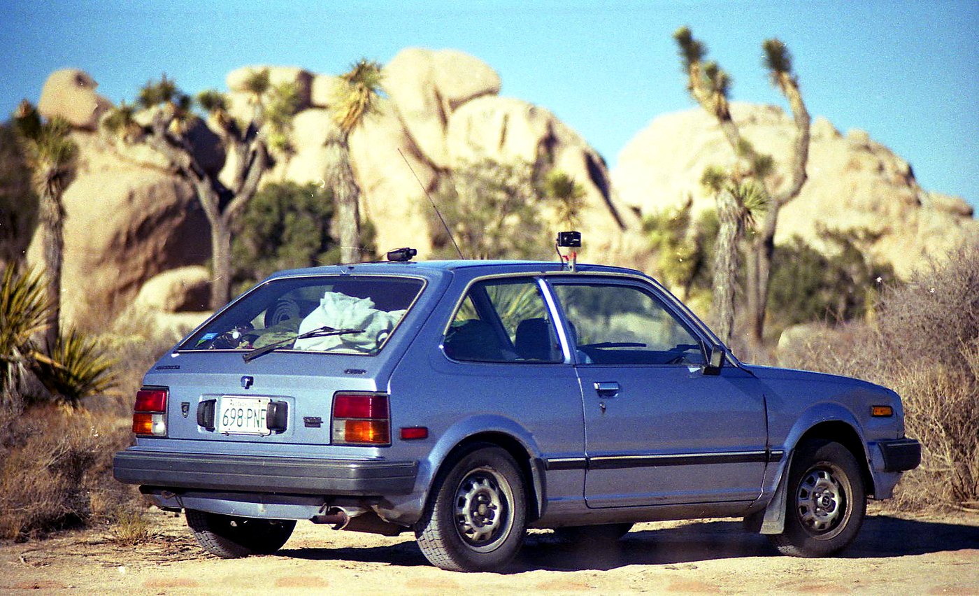 A small Honda loaded with equipment among Joshua trees and desert boulders.