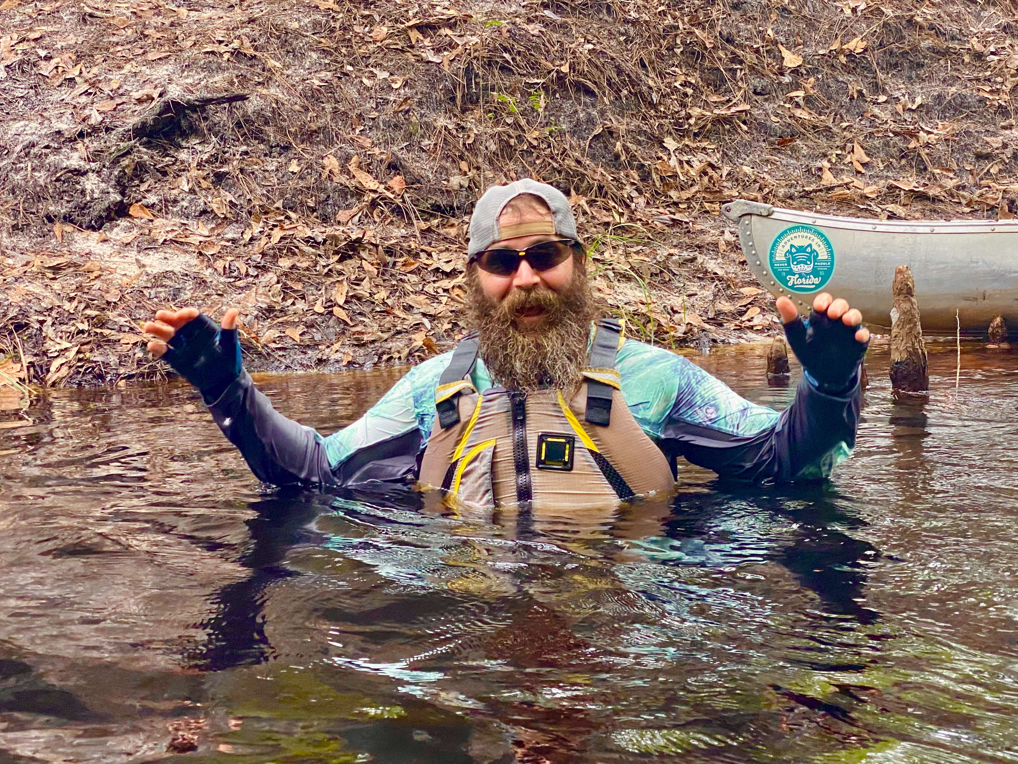 A bearded paddler standing chest-deep in the Sopchoppy River beside a canoe