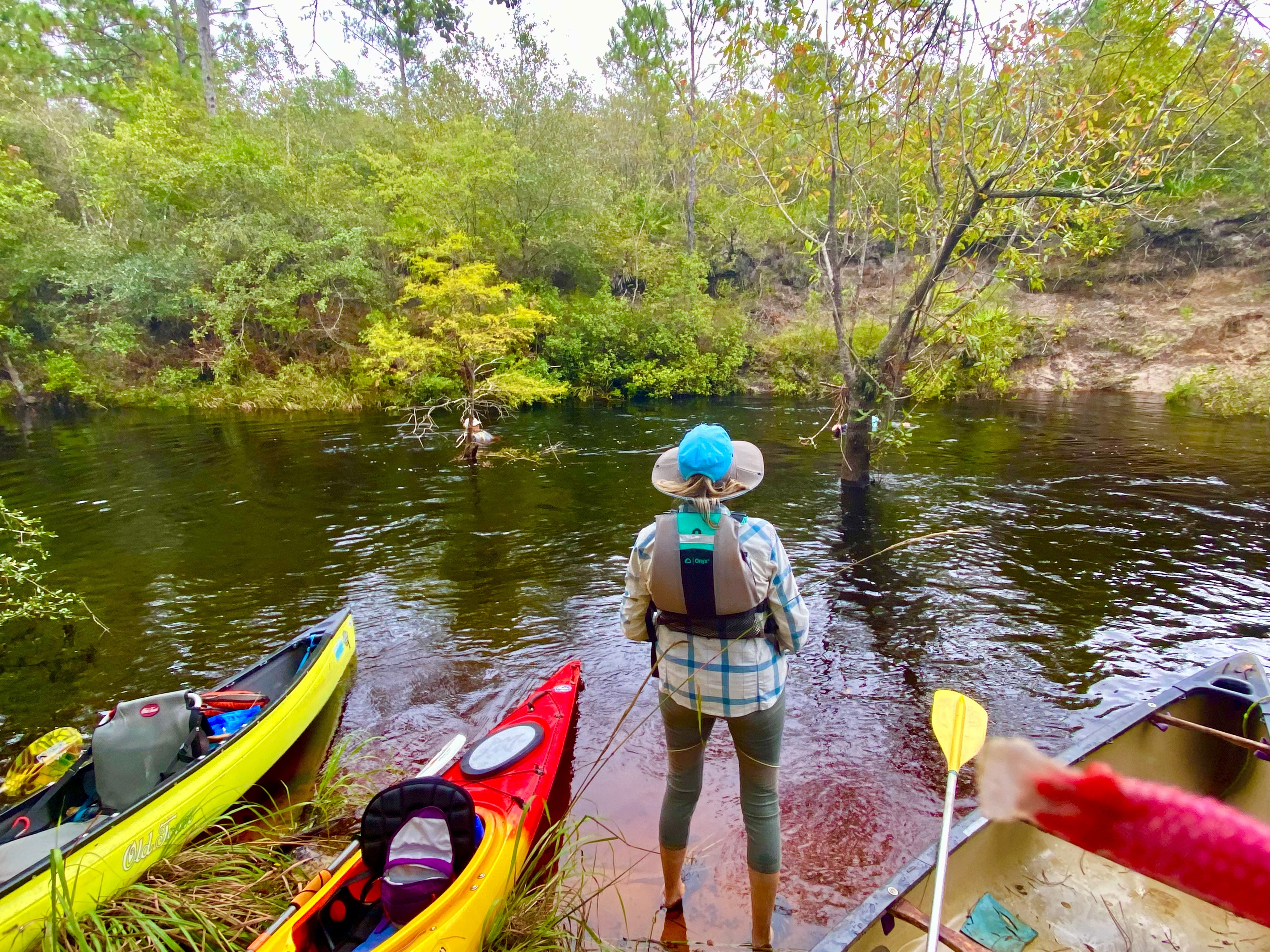 Paddlers standing beside kayaks and canoes at a wooded Sopchoppy River stop