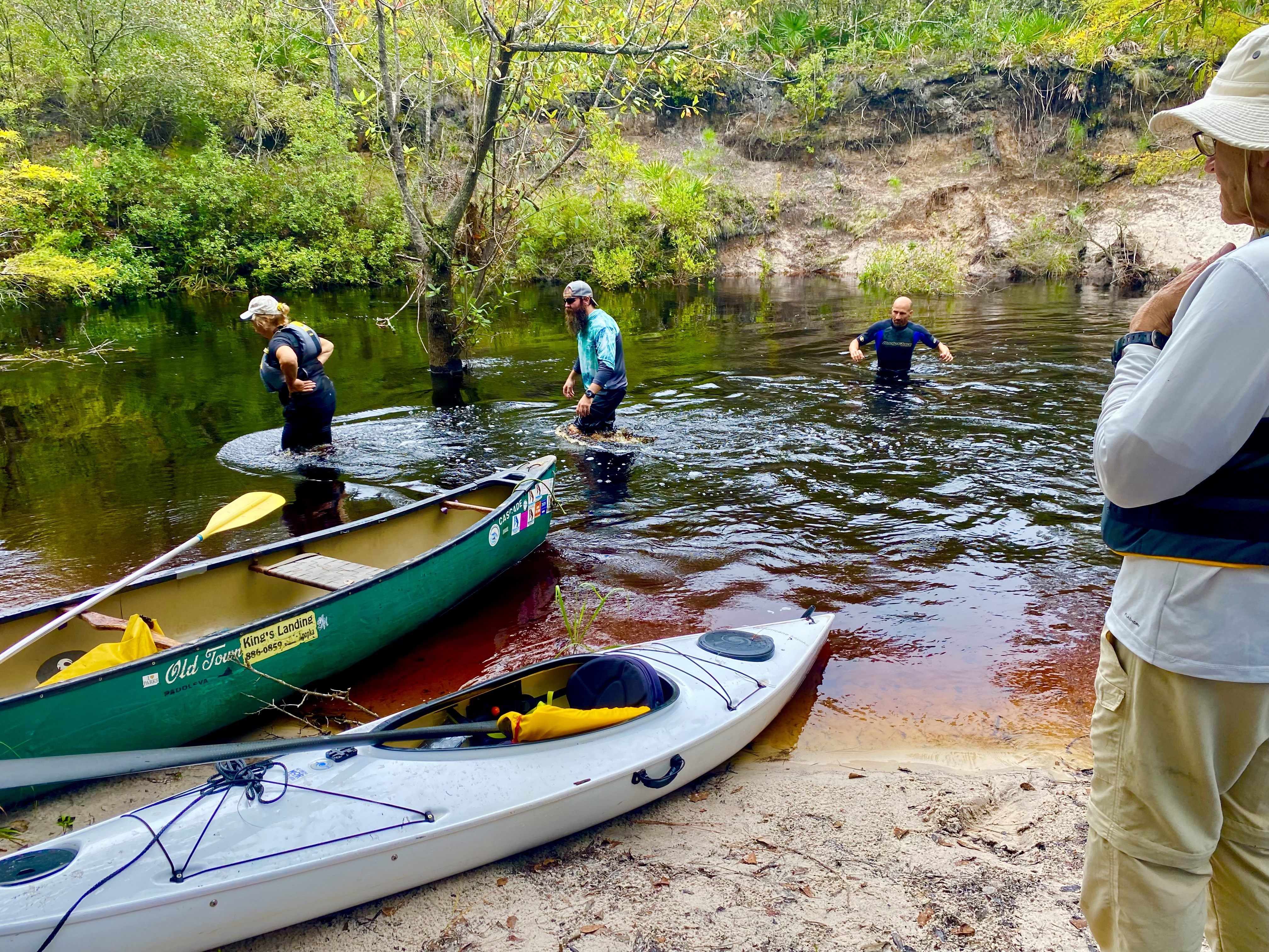 Paddlers standing in shallow water beside boats at a sandy Sopchoppy River landing