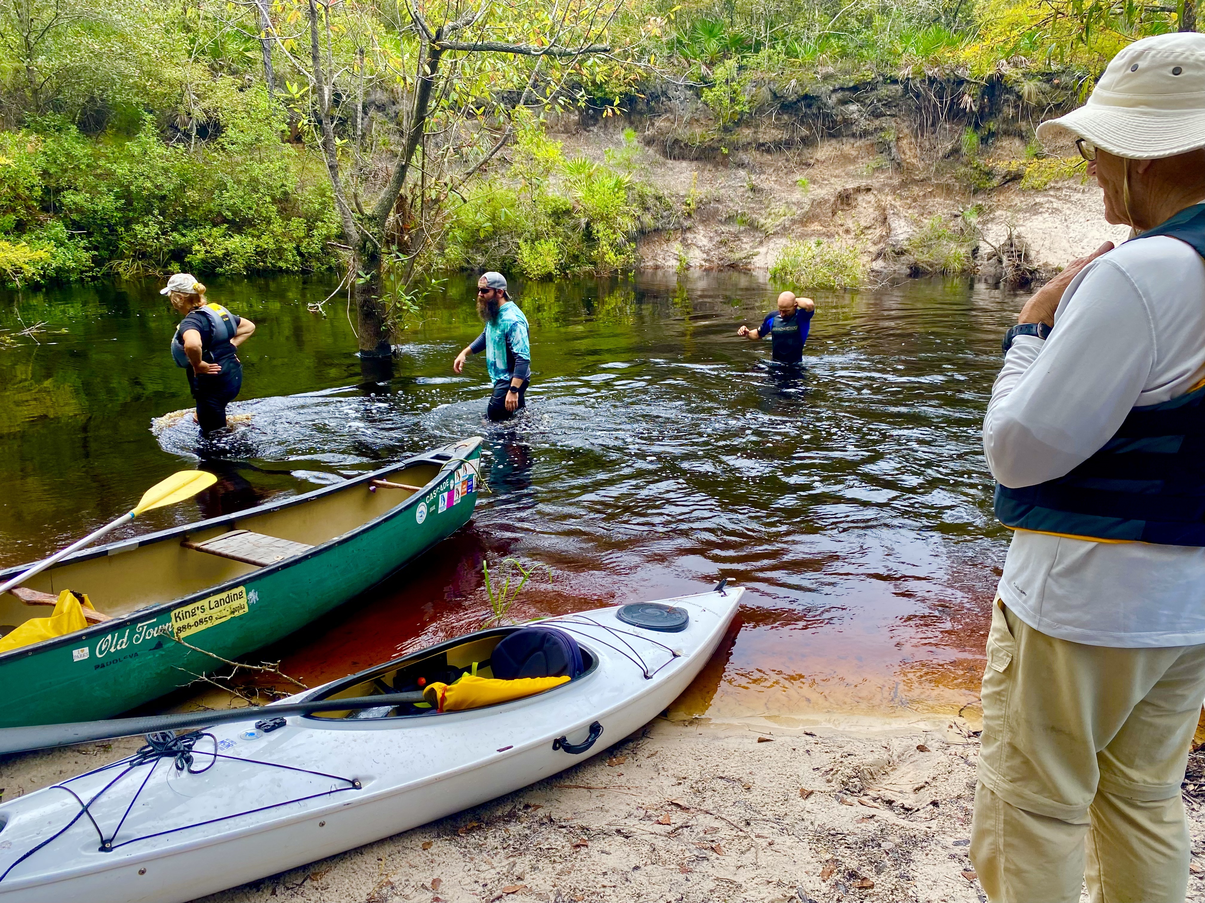 Kayaks and a canoe beside paddlers at a sandy bank on the Sopchoppy River