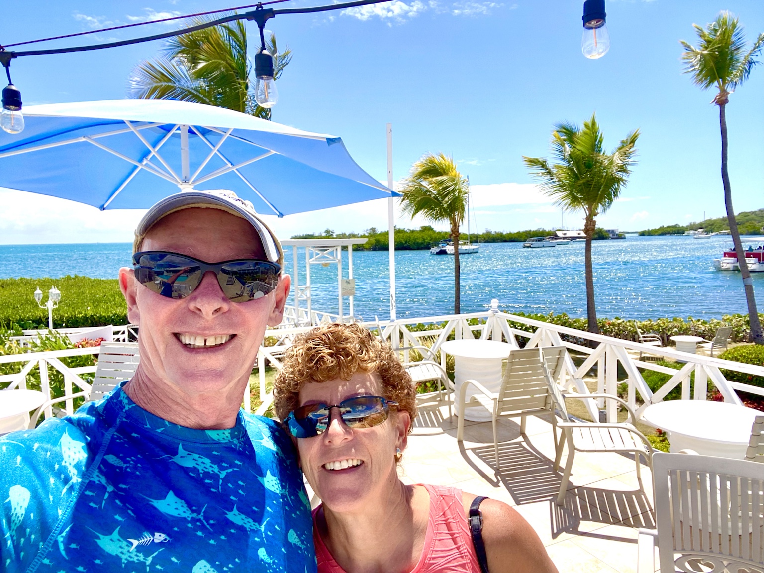 Tim and Kym smiling beside a bright tropical waterfront lined with palm trees and boats.