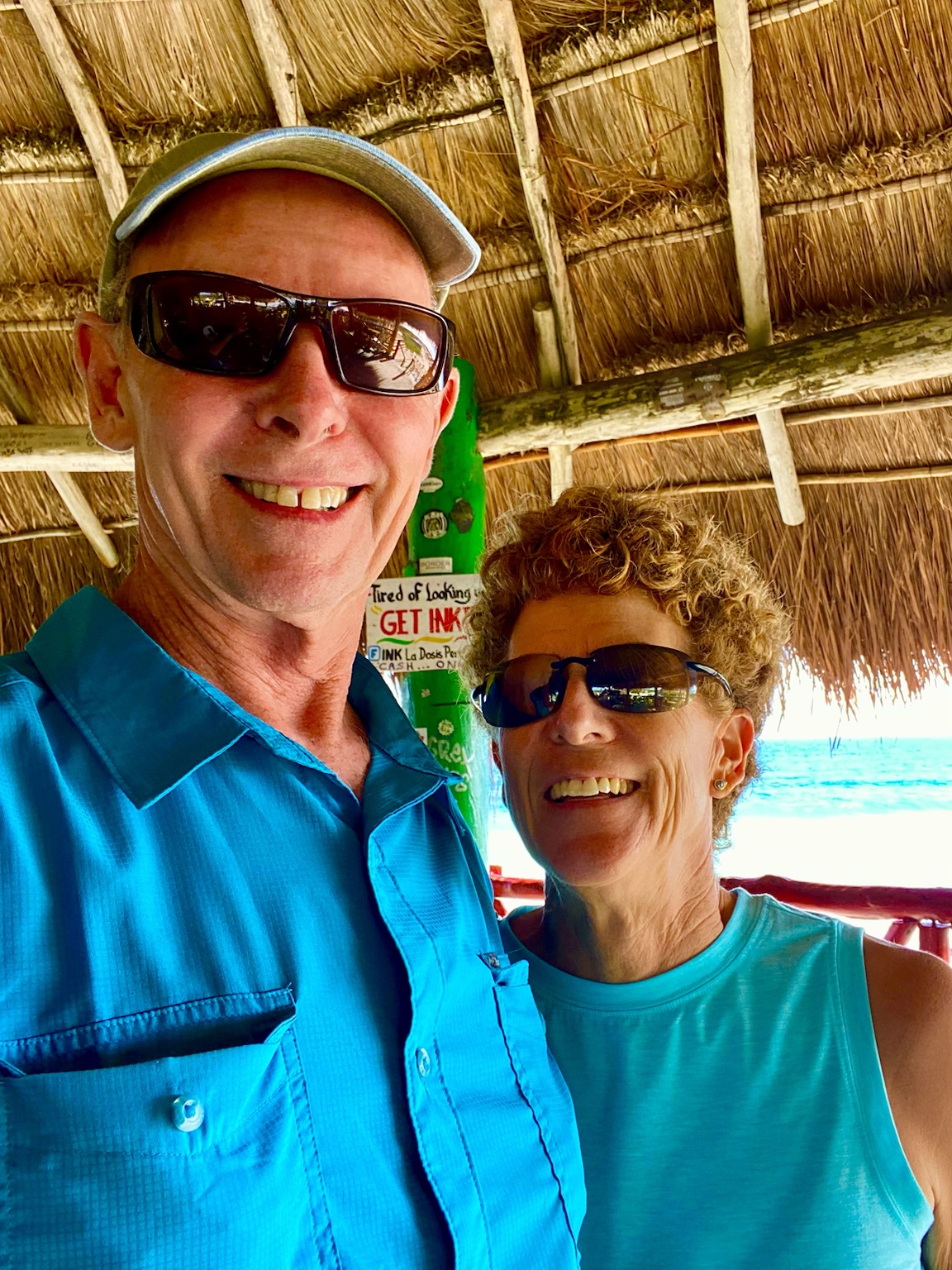 Tim and Kym beneath a thatched roof with brilliant turquoise water behind them.