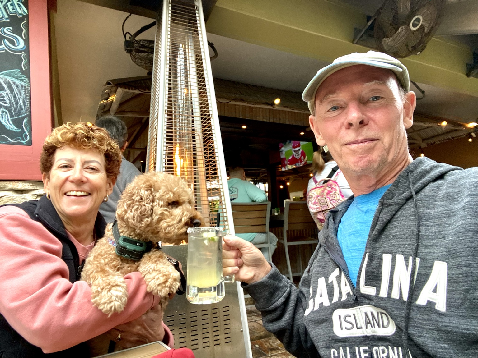 Kym holds Buddyboy while Tim raises a drink beside an outdoor patio heater.