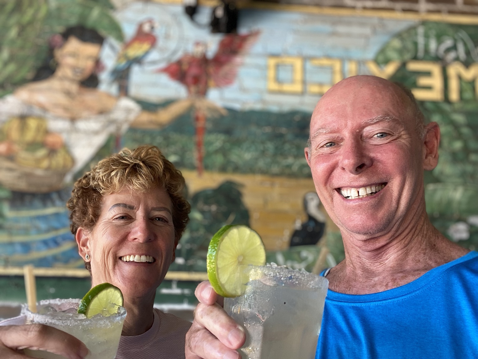 Tim and Kym toast with margaritas in front of a colorful Mexican mural.