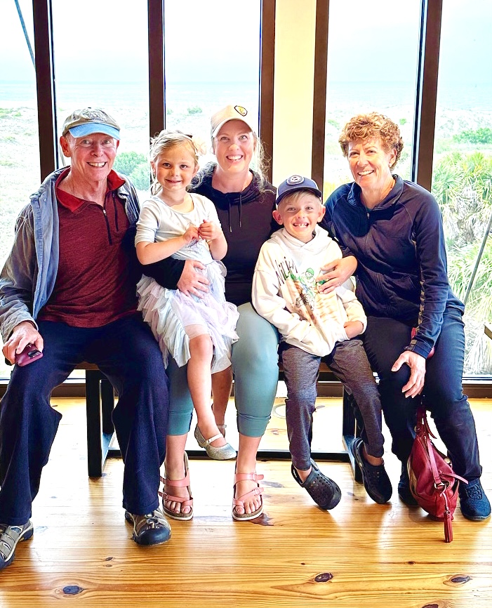 Tim and Kym with family, including two smiling children, in front of large beach windows.